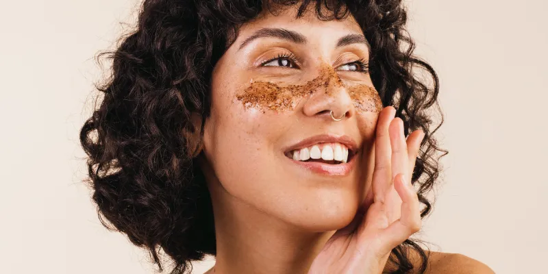 Mulher sorrindo enquanto aplica um esfoliante facial natural na área das bochechas, destacando o uso de produtos veganos e orgânicos em sua rotina de cuidados com a pele.