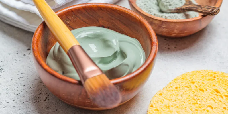 Máscara facial de argila verde em um pote de madeira com pincel aplicador, representando tratamentos de skincare naturais e veganos, ideais para purificação e cuidados com a pele.