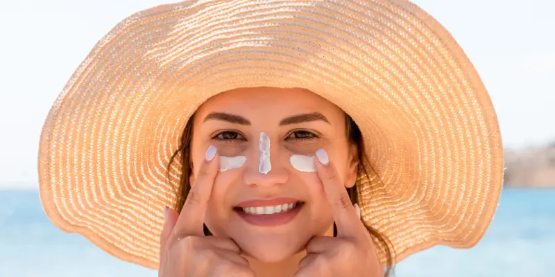 Mulher aplicando protetor solar no rosto enquanto usa um chapéu de palha, destacando a importância da proteção solar para peles sensíveis, especialmente em uma rotina de skincare.
