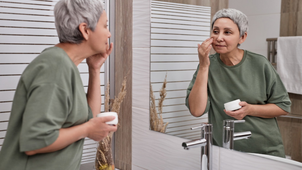 Mulher aplicando creme facial em frente ao espelho como parte de sua rotina anti-idade, cuidando da pele com foco em hidratação e prevenção de sinais de envelhecimento.