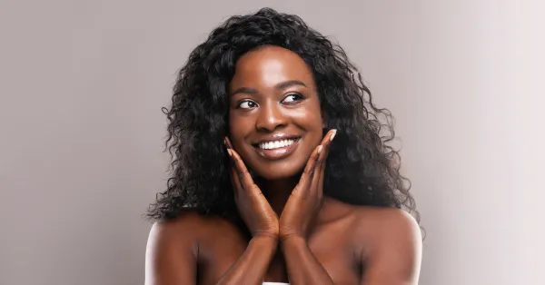 Mulher com pele radiante e sorriso confiante, tocando o rosto com as mãos, representando os benefícios de uma rotina de cuidados com a pele eficaz.