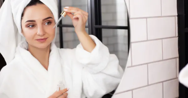 Mulher aplicando sérum no rosto em frente ao espelho, vestindo roupão e toalha na cabeça, representando uma rotina de skincare diária para cuidar da pele.