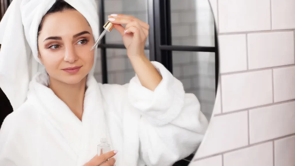 Mulher aplicando sérum no rosto em frente ao espelho, vestida com roupão e toalha na cabeça, representando uma rotina de skincare e cuidados com a pele.